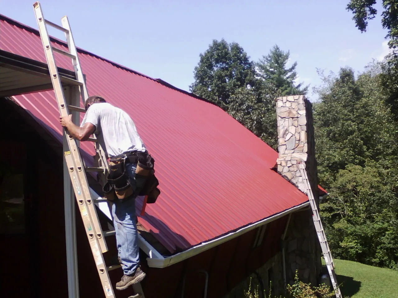 Roofer on a ladder beside a red metal roof and stone chimney — Soffit & Fascia Repair by Roofing Brothers in Willow Wood